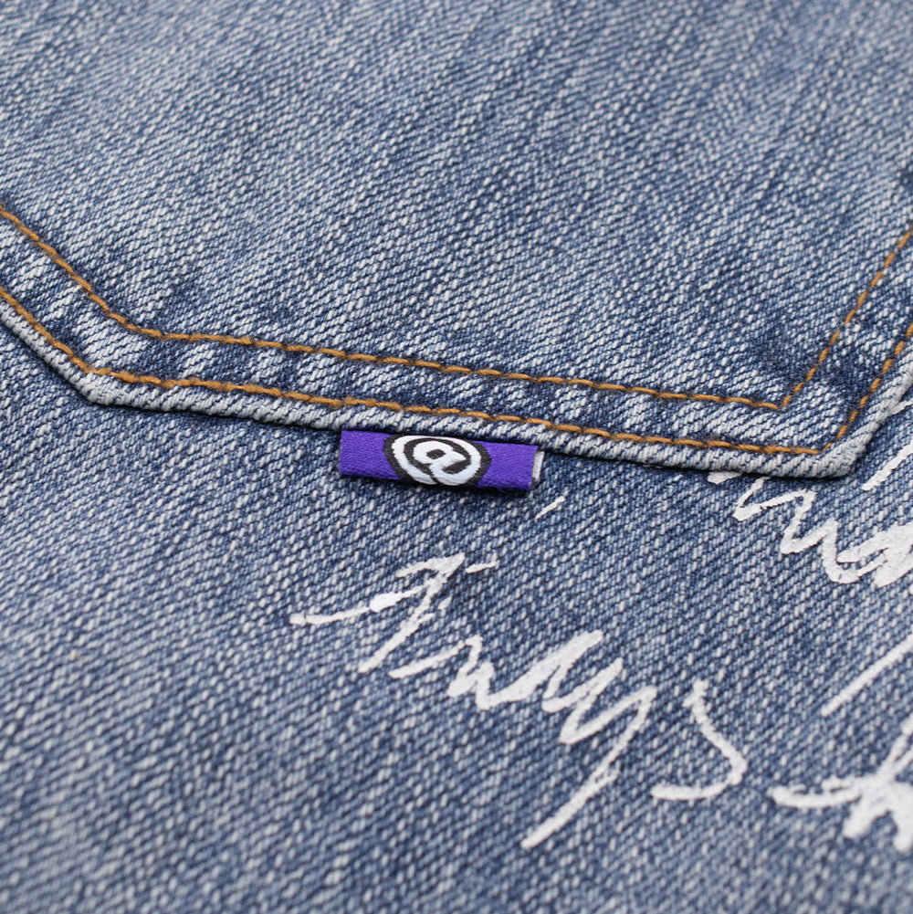 Studio macro detail of the rear pocket seam on the Always Waza Jeans, highlighting the purple woven mini flag label featuring the white circular @ logo, clean double-needle pocket topstitching, and white street-art script printing on light wash cotton denim.