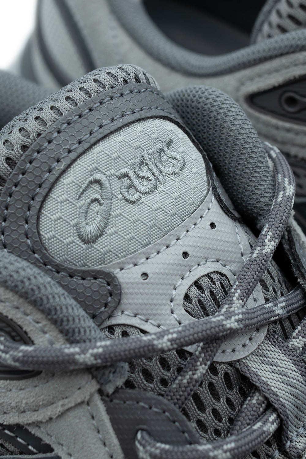 Macro close-up shot of the tongue and lace unit on the Asics Gel-NYC 2.0, detailing the hexagonal honeycomb textile tongue with embroidered cursive "Asics" lettering, perforated eyelet overlays, reflective lace loop anchor, and durable speckled grey rope laces.