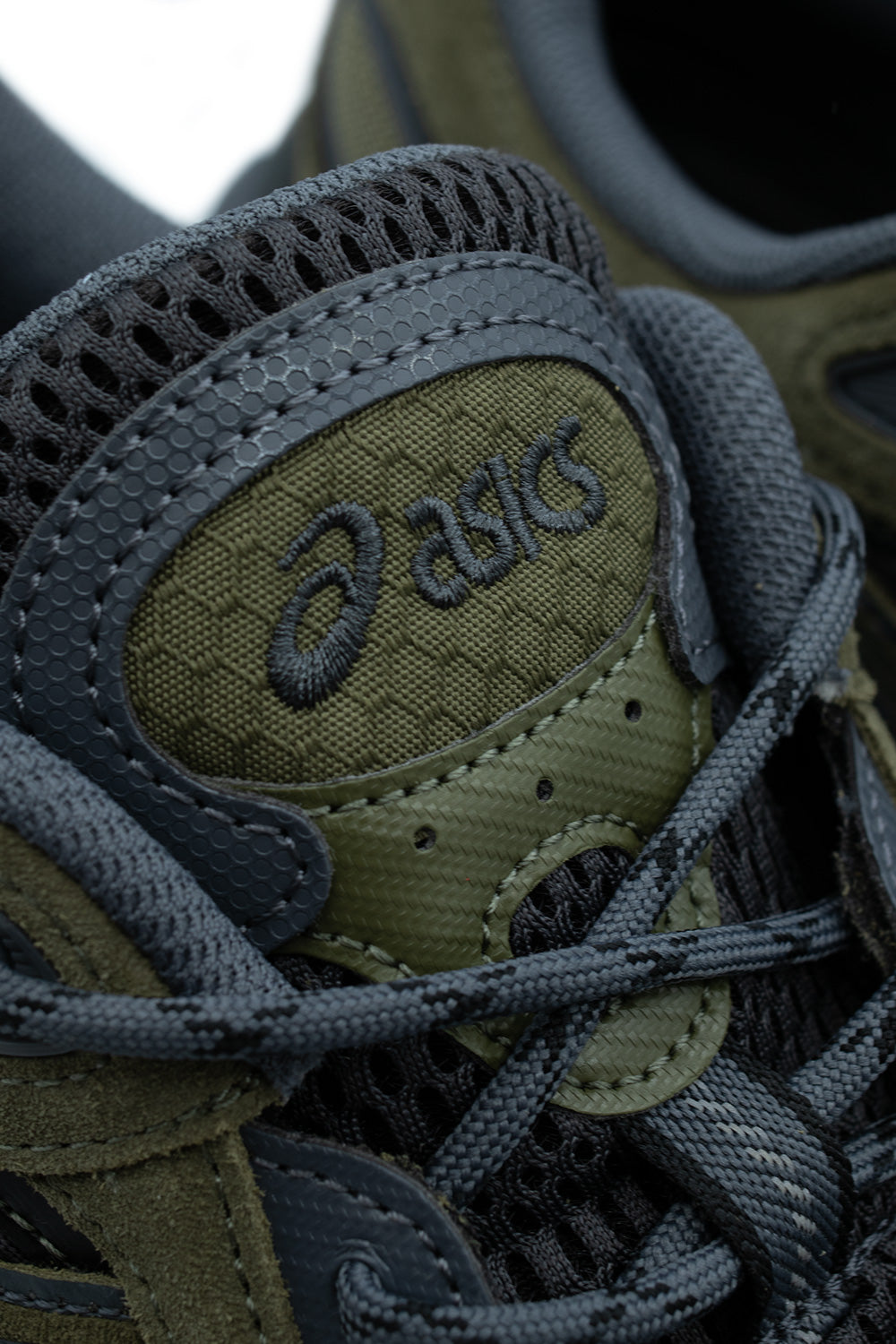 Macro close-up shot of the tongue and lacing system on the Asics Gel-NYC 2.0, detailing the hexagonal textured honeycomb olive tongue pad with dark embroidered "Asics" branding, perforated eyestay accents, and round woven graphite grey cord laces