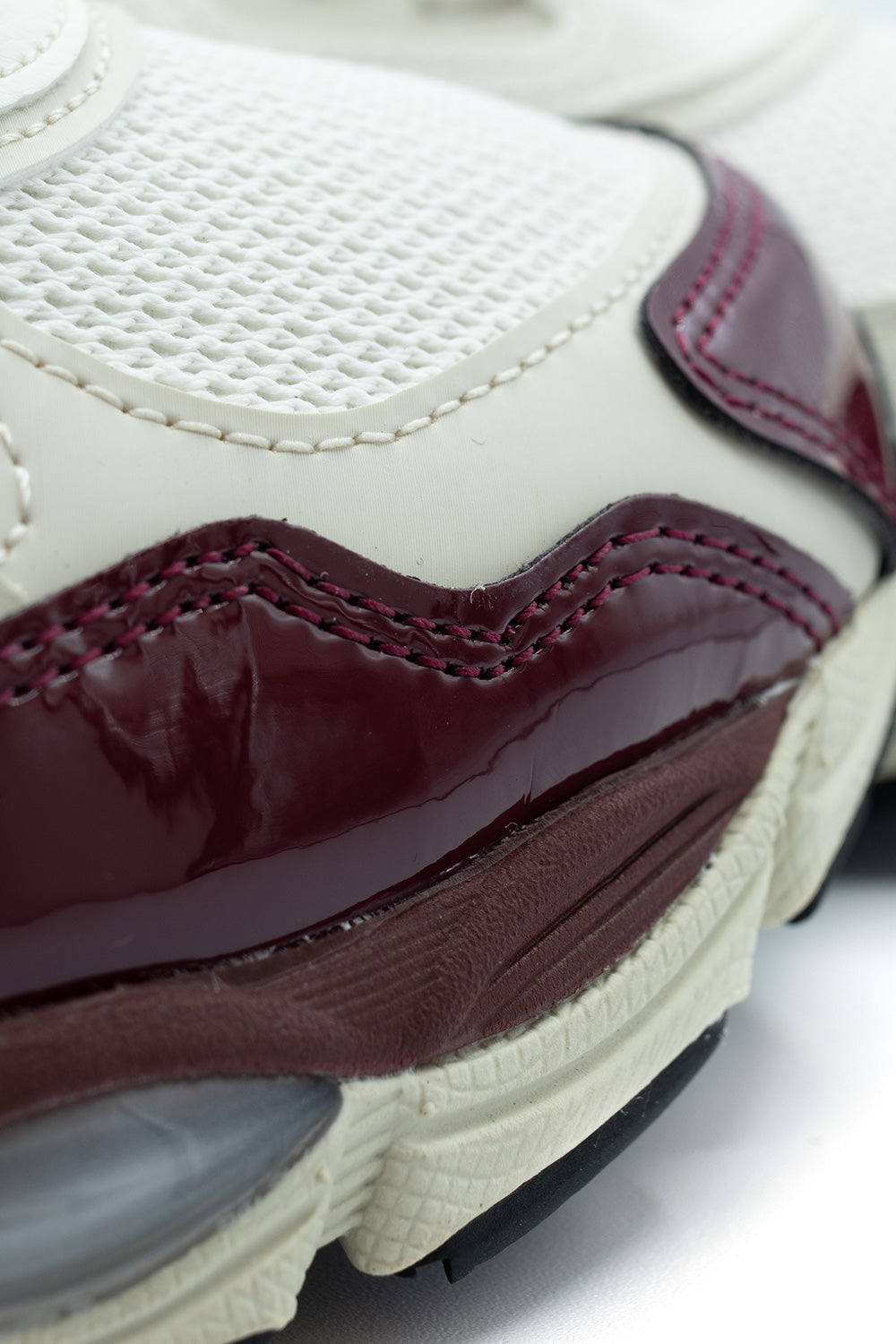 Macro detail of the forefoot construction on the Asics Gel-NYC, focusing on the double-stitched deep burgundy patent leather mudguard, tight-weave cream mesh underlay, textured midsole rim, and embedded forefoot GEL cushioning unit