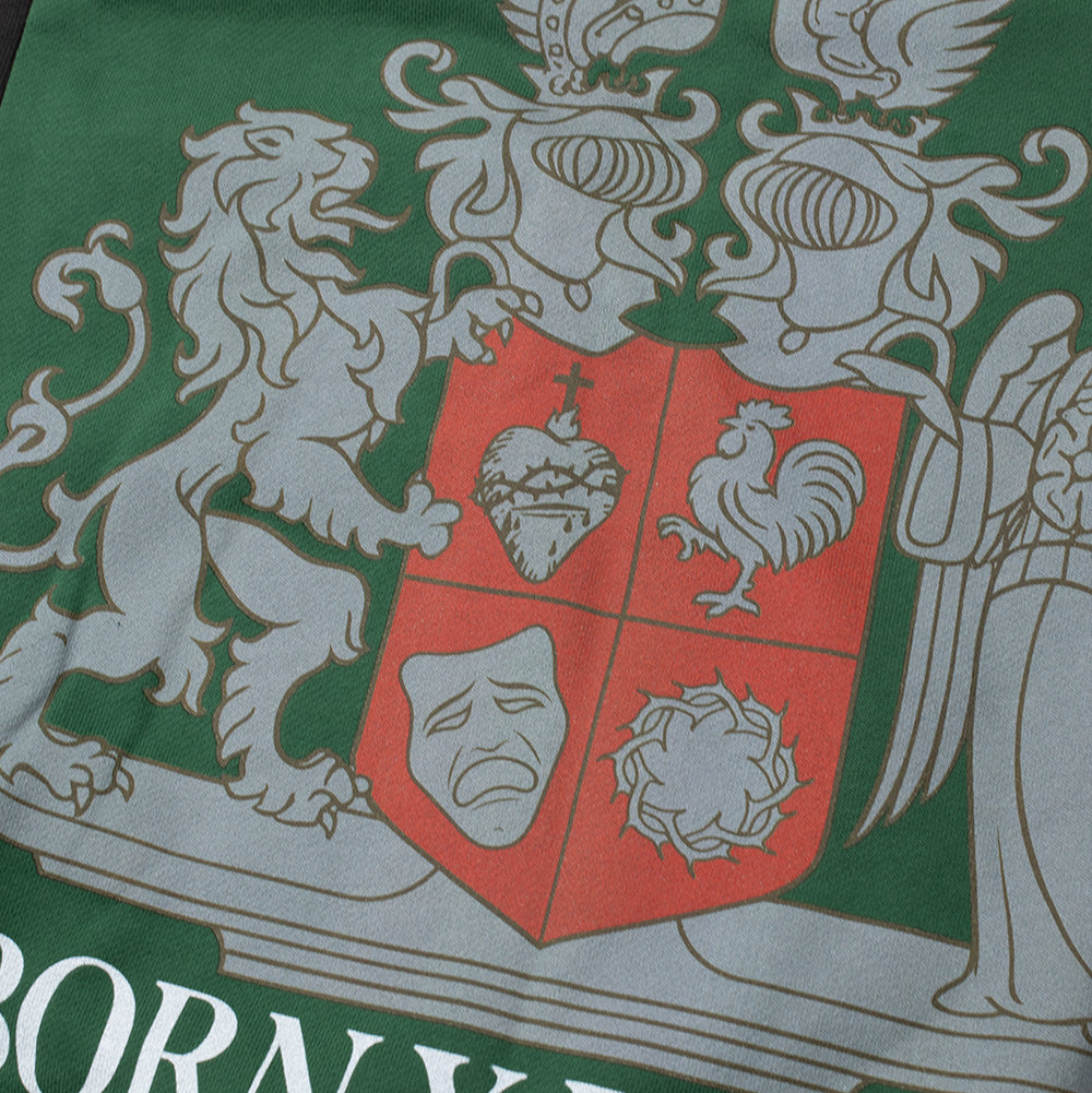 Macro close-up shot of the lower heraldic emblem on the green fleece side of the Born X Raised Mashup Hoodie, detailing the intricate silver-grey and red coat of arms depicting a rampant lion, armored knights, Sacred Heart, rooster, comedy mask, and crown of thorns above "BORN X RAISED" block text.