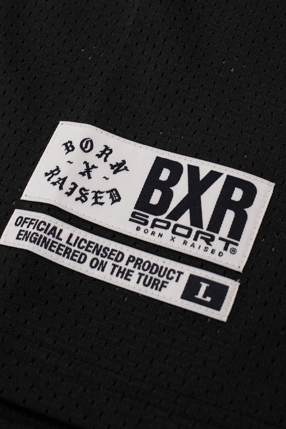 Detailed macro view of the lower hem woven jock tag label reading BXR Sport Born X Raised Official Licensed Product on the black mesh fabric.