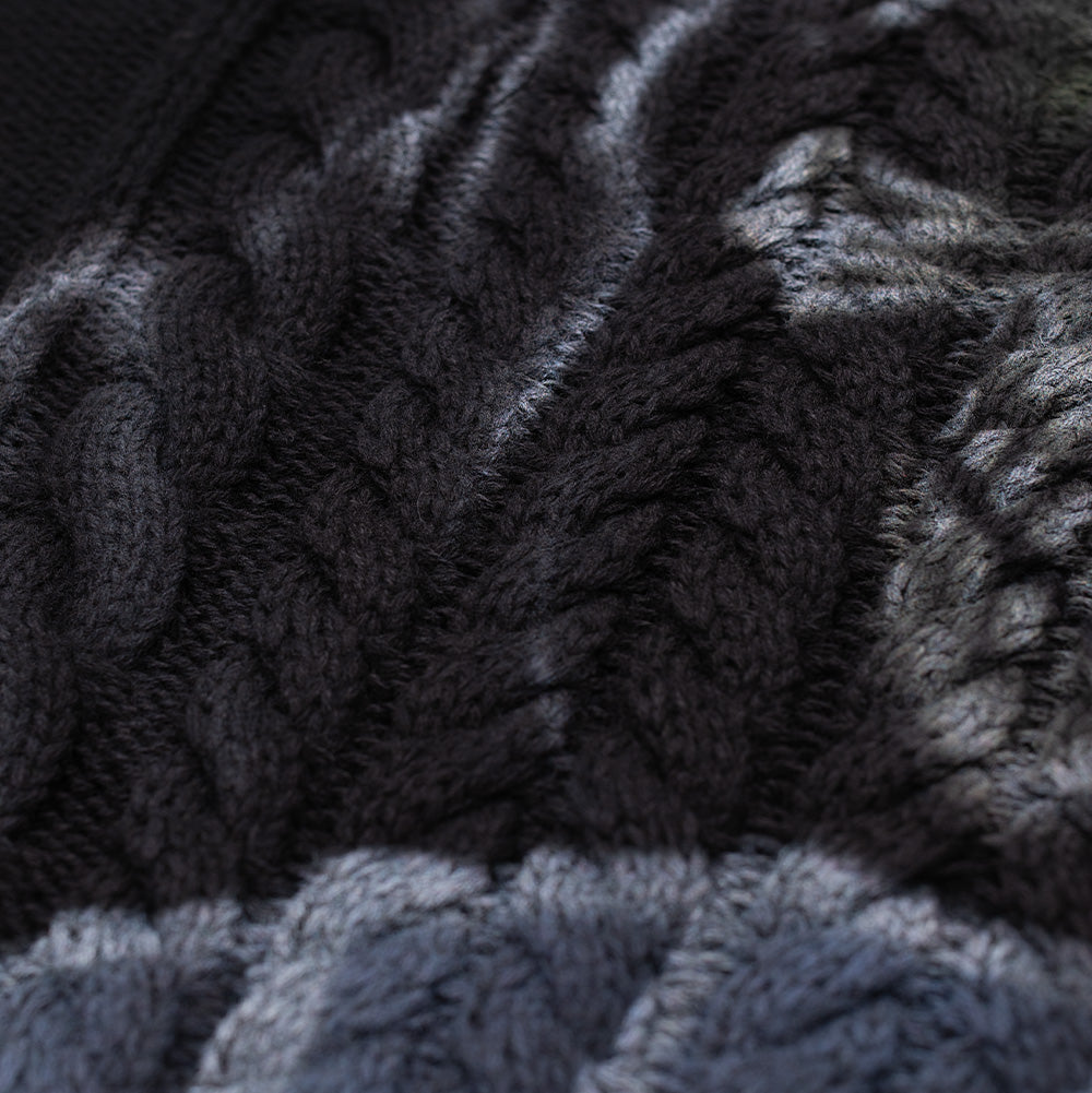 Extreme macro close-up of the knitted fabric on the Born X Raised Two Pair Sweater, detailing the heavy gauge black yarn woven into dimensional rope cable ropes, textured diamond purl stitches, and the smooth shading of the front artwork.