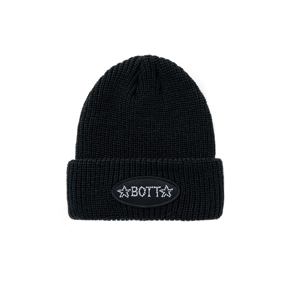 Front flat lay studio view of the BoTT Bones Cuff Beanie in solid black, showcasing a chunky ribbed-knit acrylic fisherman beanie with a wide folded cuff, centered with an oval black woven twill patch featuring white embroidered "BoTT" typography and flanking stars composed of illustrated human bones.