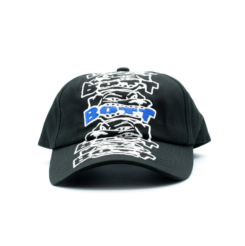 Front perspective studio view of the BoTT Bottweiler 6 Panel Cap in durable black cotton twill, showcasing an unstructured skate cap with a curved brim and an oversized, vertically extending embroidery of stacked cartoon rottweiler faces in white thread with vibrant royal blue "BoTT" block lettering in the center.
