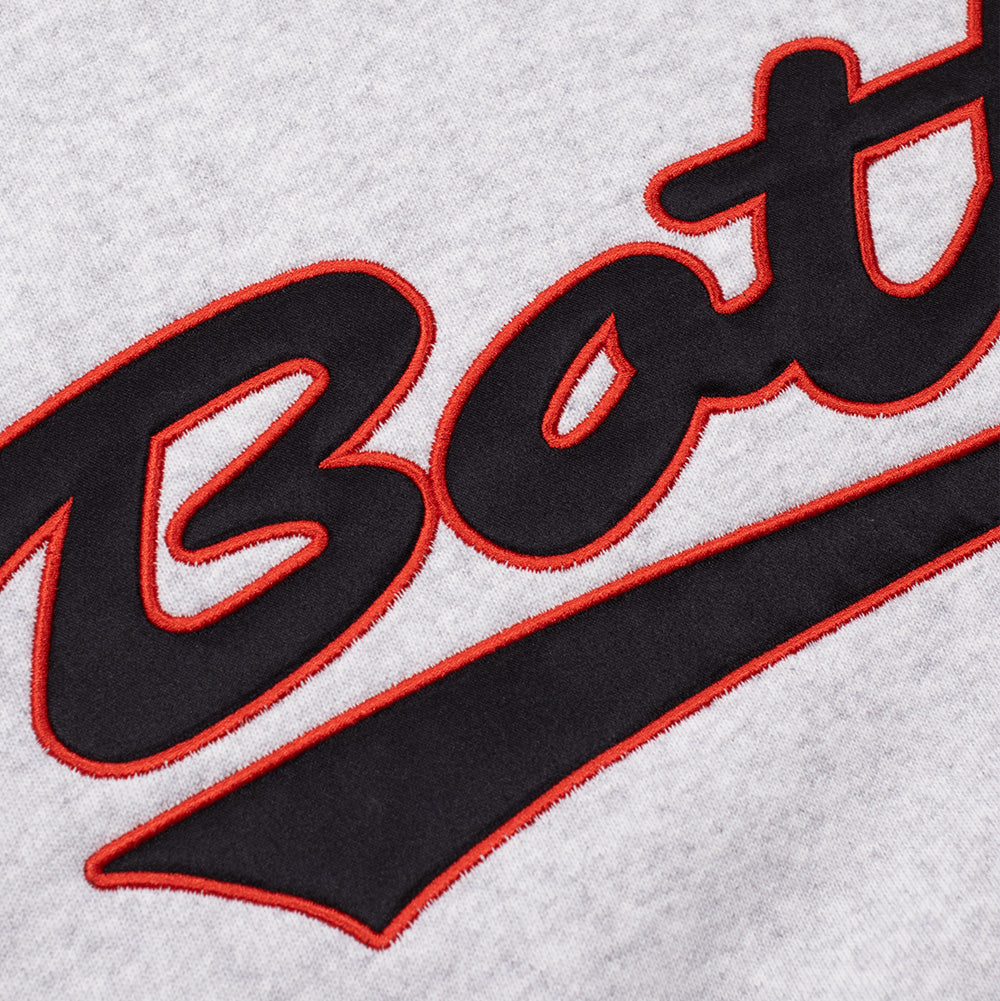 Detailed macro perspective of the chest embroidery on the ash grey BoTT Script Logo Hoodie, highlighting the tactile black twill appliqué lettering tightly secured by dense red satin-stitch edge embroidery to form the classic baseball-inspired "BoTT" signature script.
