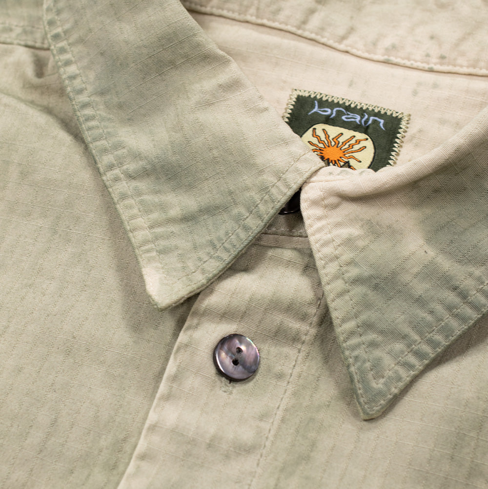 Detailed macro perspective of the pointed collar and top button placket on the Brain Dead Ripstop Cropped Shirt Jacket, displaying the woven green interior neck tag featuring the sun motif and white "brain" lettering, beside a dark iridescent mother-of-pearl button sewn into the textured, crosshatch ripstop grid weave.