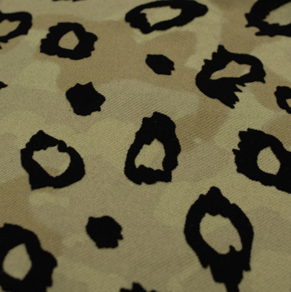 Extreme macro texture perspective of the Brain Dead canvas fabric, detailing the mottled camouflage color variations in beige and olive, overlaid with distinctly raised, velvety black flocking creating the organic edges of the leopard animal spot motif.