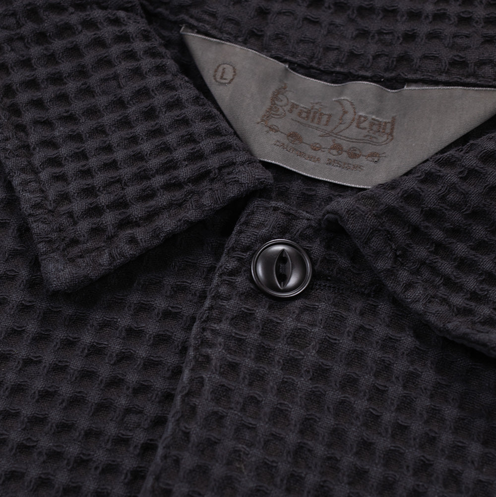 Detailed macro studio close-up of the collar neckline on the Brain Dead Waffle Button Front Shirt, highlighting the grey satin interior brand tag with embroidered gothic script and California Designs text, beside the thick three-dimensional square waffle grid weave and top cat-eye style black resin button.