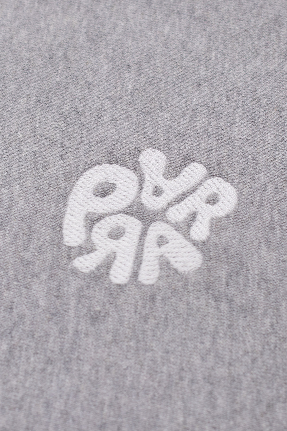 Macro close-up shot of the left chest embroidery on the heather grey By Parra crewneck sweater, detailing the textured white satin-stitch thread forming the rounded, balloon-style typography arranged in a circular Parra brand motif against the brushed cotton blend fleece.