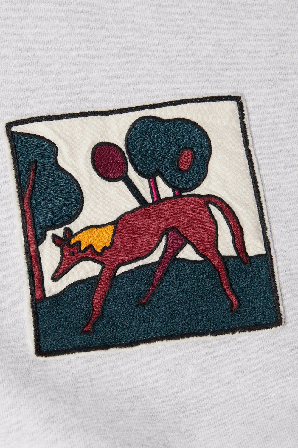 Close-up detail of the sewn-on chest patch on the By Parra sweater, displaying Pieter Janssen's signature surreal illustration of a red hound-like creature in a stylized landscape with trees on a cream fabric background.