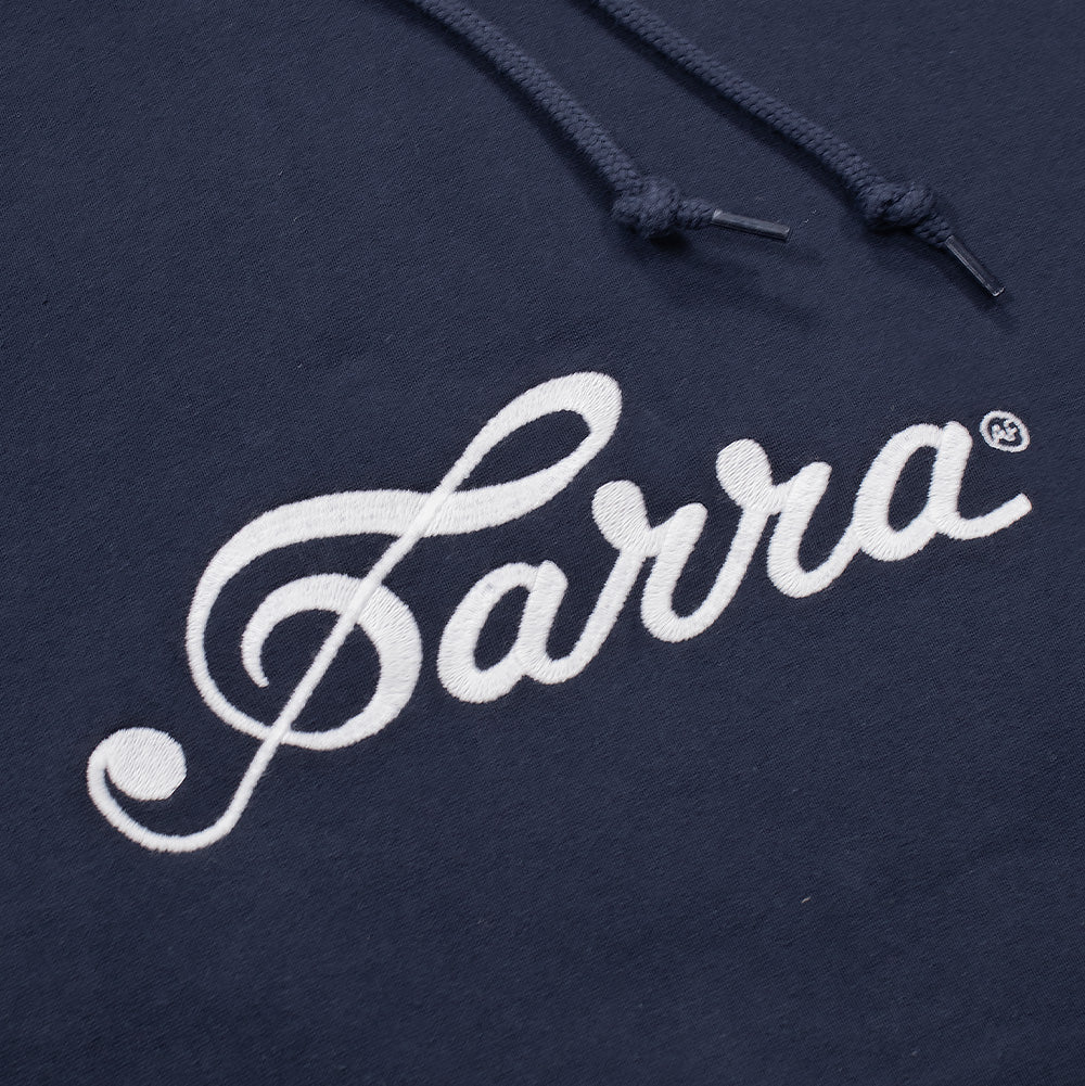 Detailed macro studio close-up of the chest embroidery on the By Parra Musical Chairs Hoodie, highlighting the high-density white satin-stitch thread forming the flowing cursive "Parra" signature script, knotted navy blue hood drawcords with plastic aglets, and the smooth texture of the dark navy cotton jersey knit fabric.