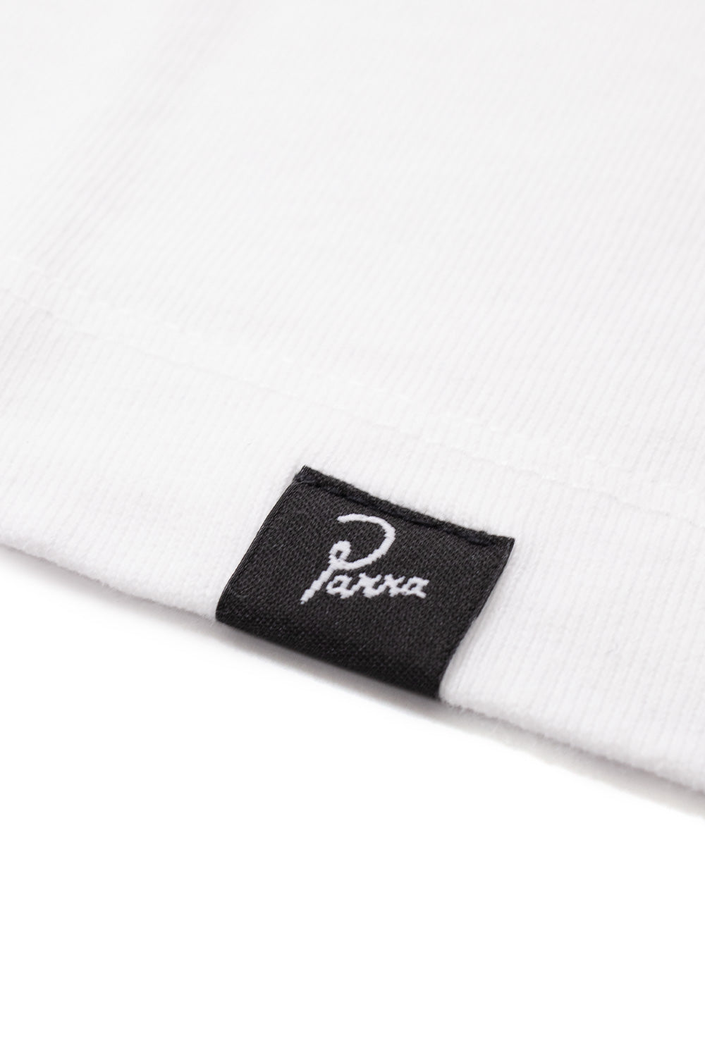 Extreme close-up view of the lower hemline on the white By Parra Not Another Boat T-Shirt, displaying the premium textured 240g cotton knit jersey, parallel twin-needle stitch finishing, and folded black woven seam tag with white embroidered Parra script.