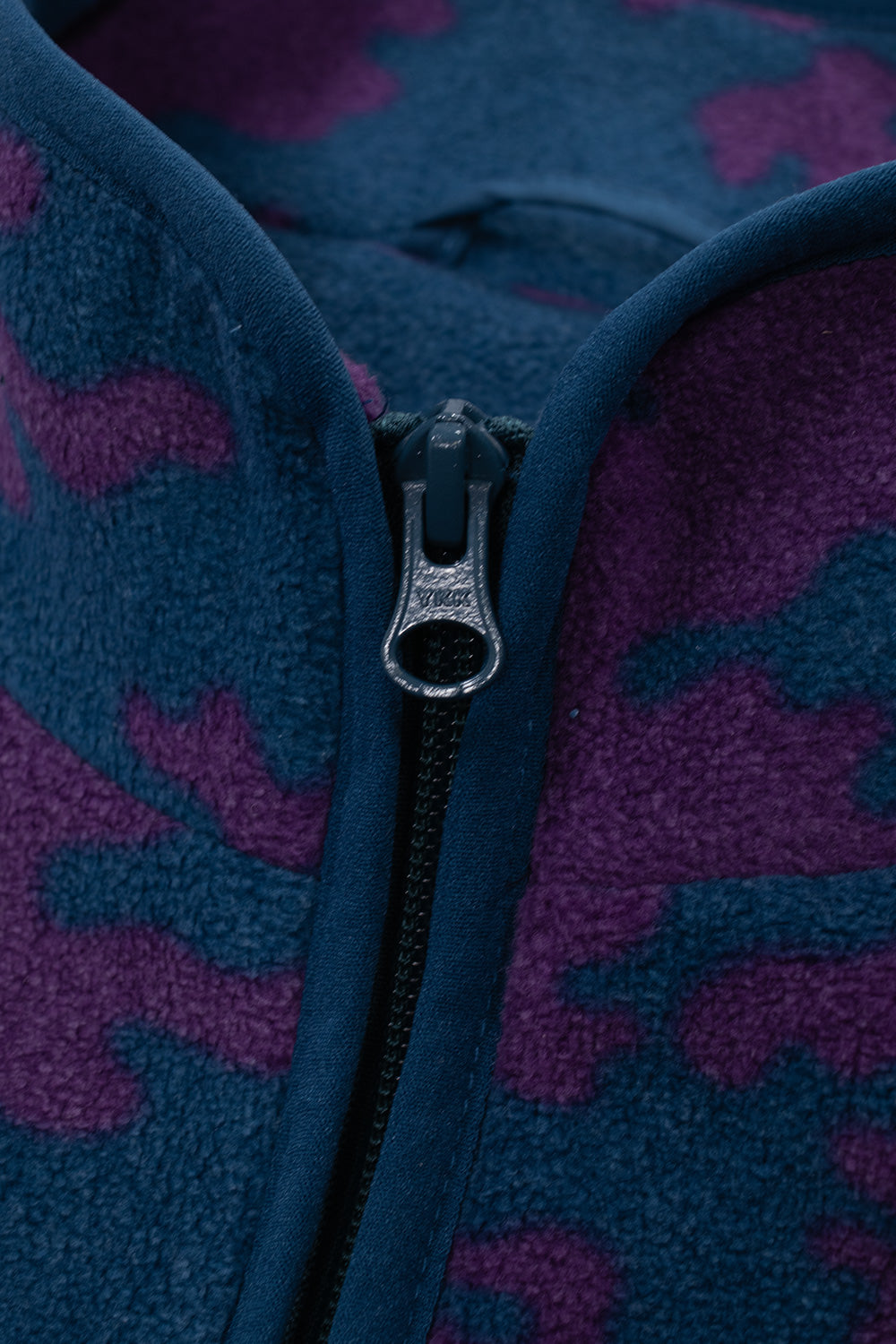 Macro close-up shot of the front neckline on the By Parra fleece, highlighting the blue YKK zipper pull, fabric zipper garage, structured stand-up collar edge, and plush purple and blue fleece fibers.