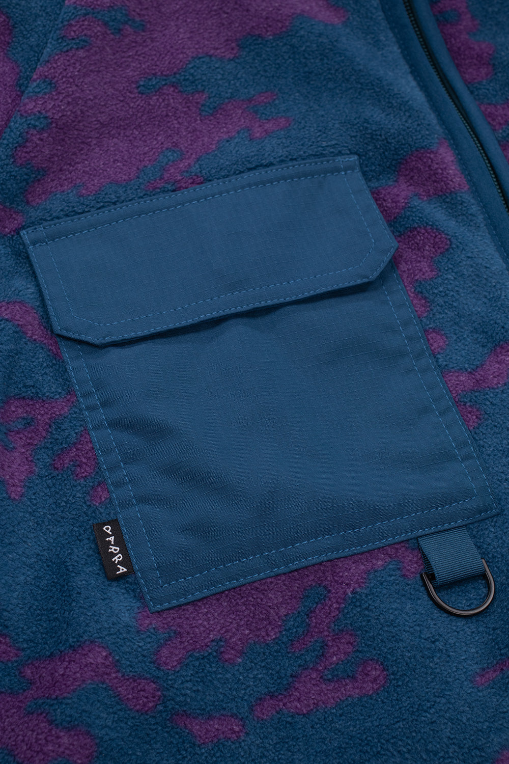 Detailed shot of the right chest ripstop flap pocket on the By Parra fleece jacket, showcasing the matte black tactical D-ring hanging from the bottom edge and the small black woven Parra label stitched into the side seam.