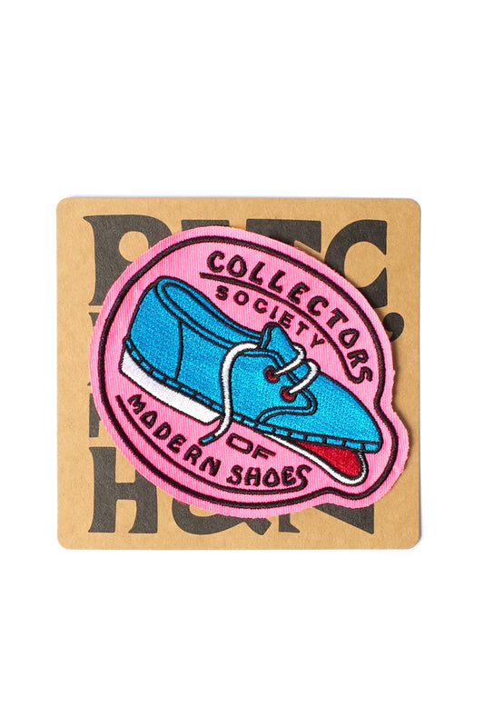 Studio shot of the By Parra Shoe Collector Embroidered Patch displayed on its brown kraft paper backing card, featuring an oval pink twill badge measuring 11 cm by 9 cm with a black stitched merrowed border, centered around a bright cyan blue retro deck shoe illustration with a red mouth opening and arched black lettering reading "COLLECTORS SOCIETY OF MODERN SHOES".