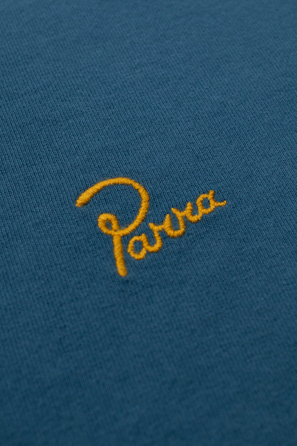 Detailed close-up view of the yellow cursive Parra brand logo embroidery stitched on the heavy teal blue cotton fabric of the By Parra Signature tee.
