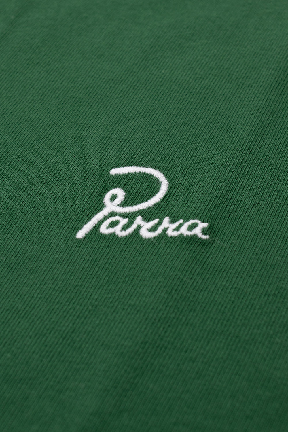 Macro close-up shot of the left chest on the green By Parra Signature T-Shirt, displaying the fine white satin-stitch embroidered cursive Parra script logo against the densely knit, pre-washed green cotton fabric.