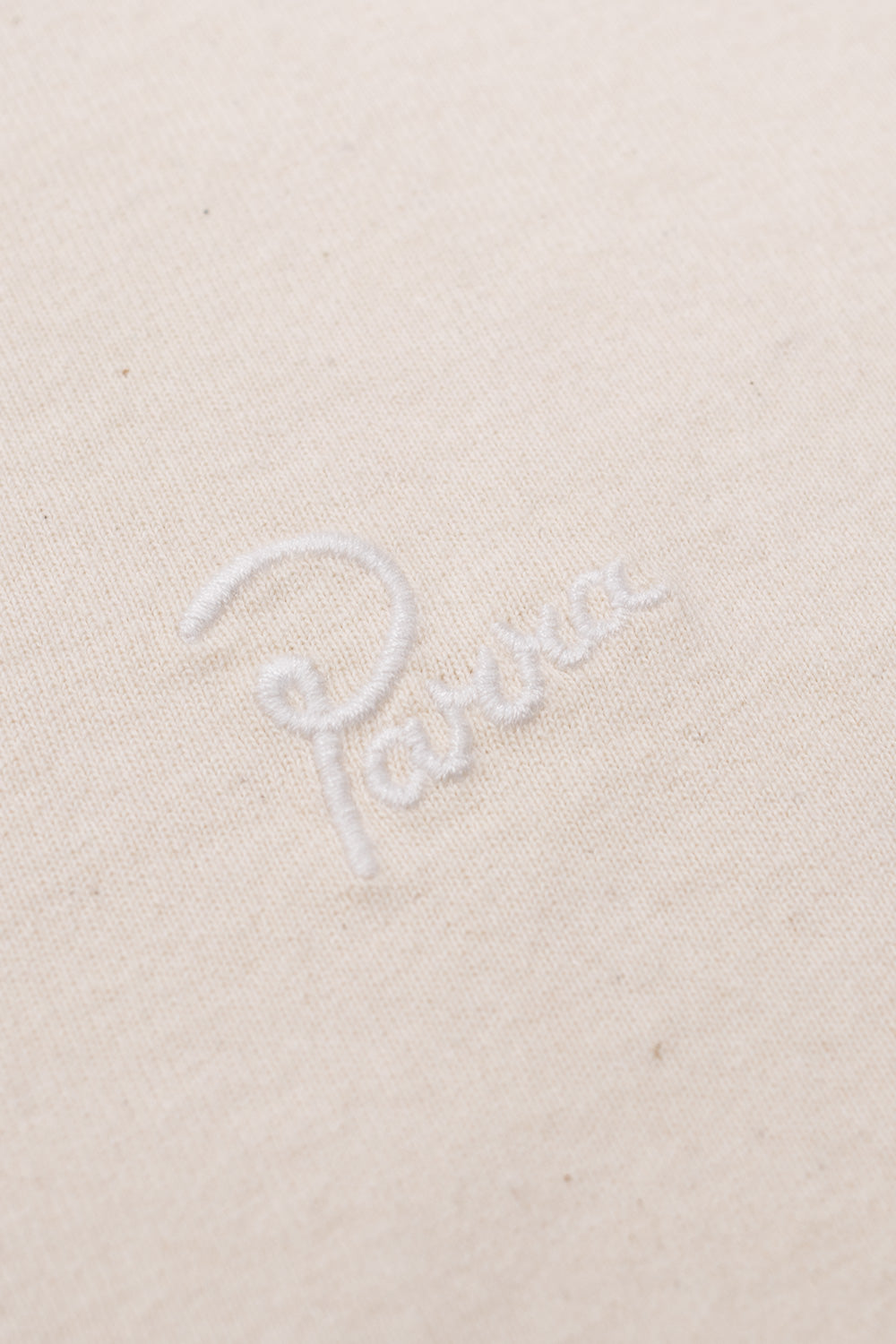 Detailed close-up shot of the white cursive Parra brand logo embroidery stitched on the heavy natural cream cotton fabric of the By Parra Signature tee.