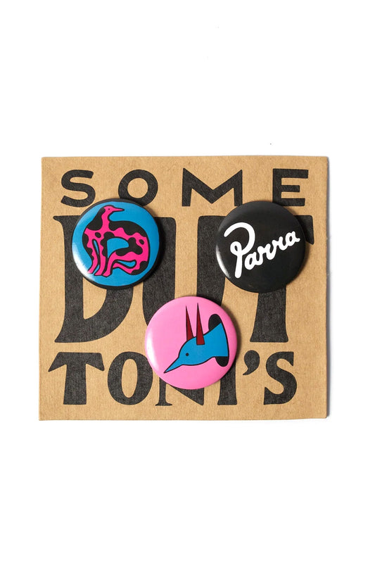 Front studio view of the By Parra Some Butonis Pin-Back Set pinned to a brown kraft cardboard backing card printed with bold black "SOME BUTTON'S" typography, displaying three 4 cm round button pins: a bright blue pin featuring a pink and black camouflage bird figure, a solid black pin with white cursive Parra script logo, and a pink pin with a blue horned bird character profile.