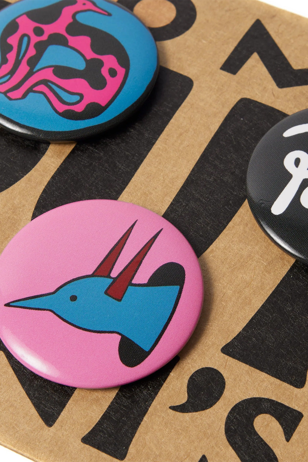 Angled macro close-up shot of the By Parra Some Butonis round button badges on the kraft paper packaging, detailing the vibrant glossy finish of the pink pin depicting a stylized light blue bird with horns and the bright blue pin printed with a pink patterned avian silhouette.