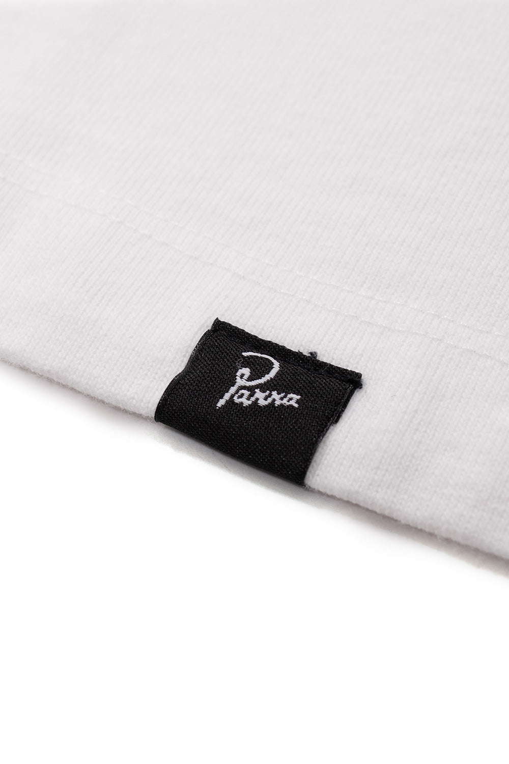 Macro view of the bottom hem on the white 100% cotton By Parra shirt, displaying a small black woven logo flag tag embroidered with white script Parra branding.