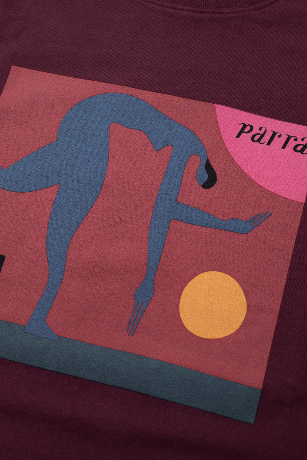 Macro close-up shot of the graphic chest screen print on the dark red By Parra Tripped Up T-Shirt, showcasing the soft water-based ink artwork of a contorted blue human hybrid character with elongated fork-like hands, a yellow sphere, pink backdrop accent, and black cursive Parra typography.