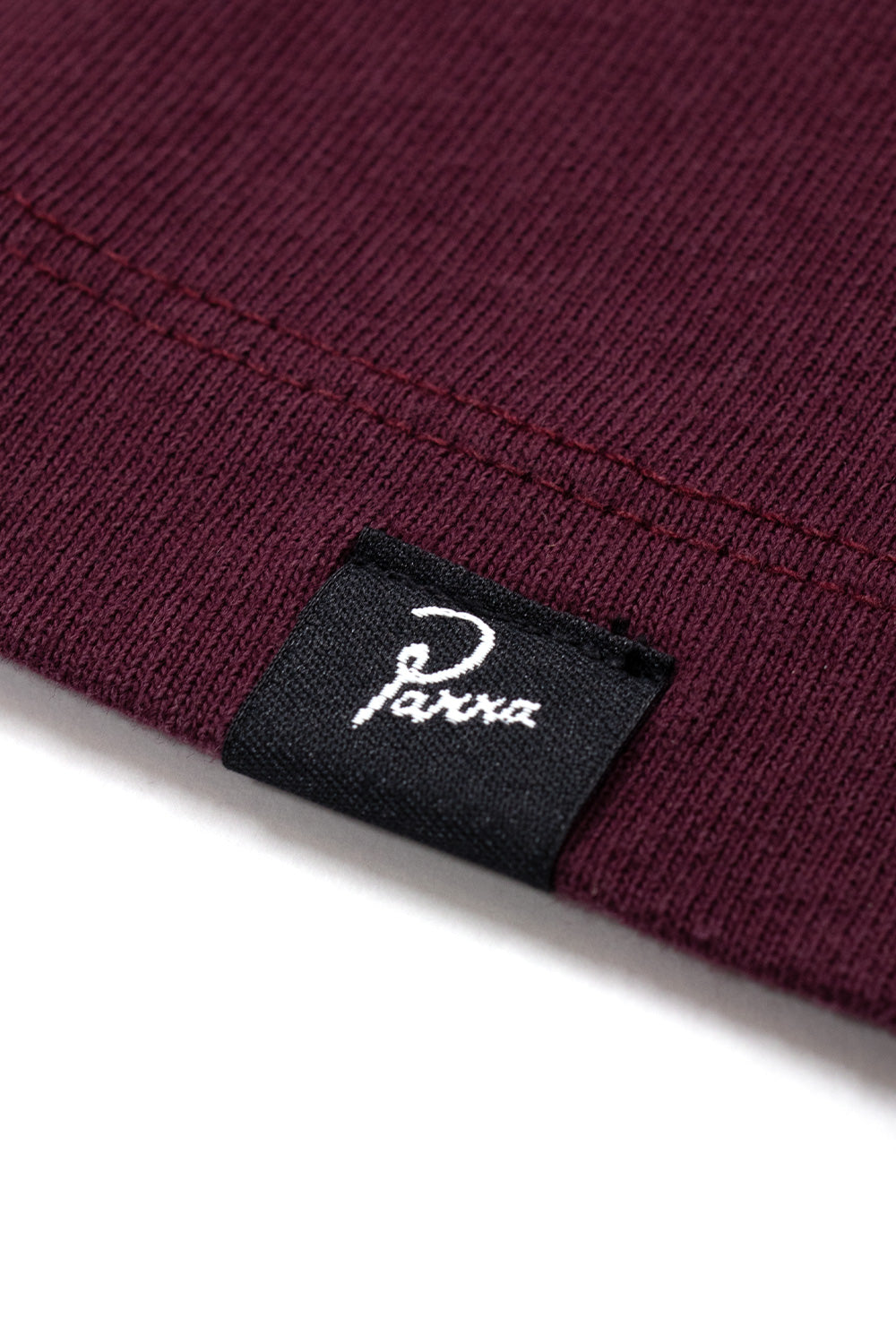 Extreme close-up detail shot of the bottom hem on the burgundy dark red By Parra T-Shirt, displaying the dense 240g knit cotton jersey, parallel twin-needle hem stitching, and folded black woven seam tag with white embroidered cursive Parra logo.