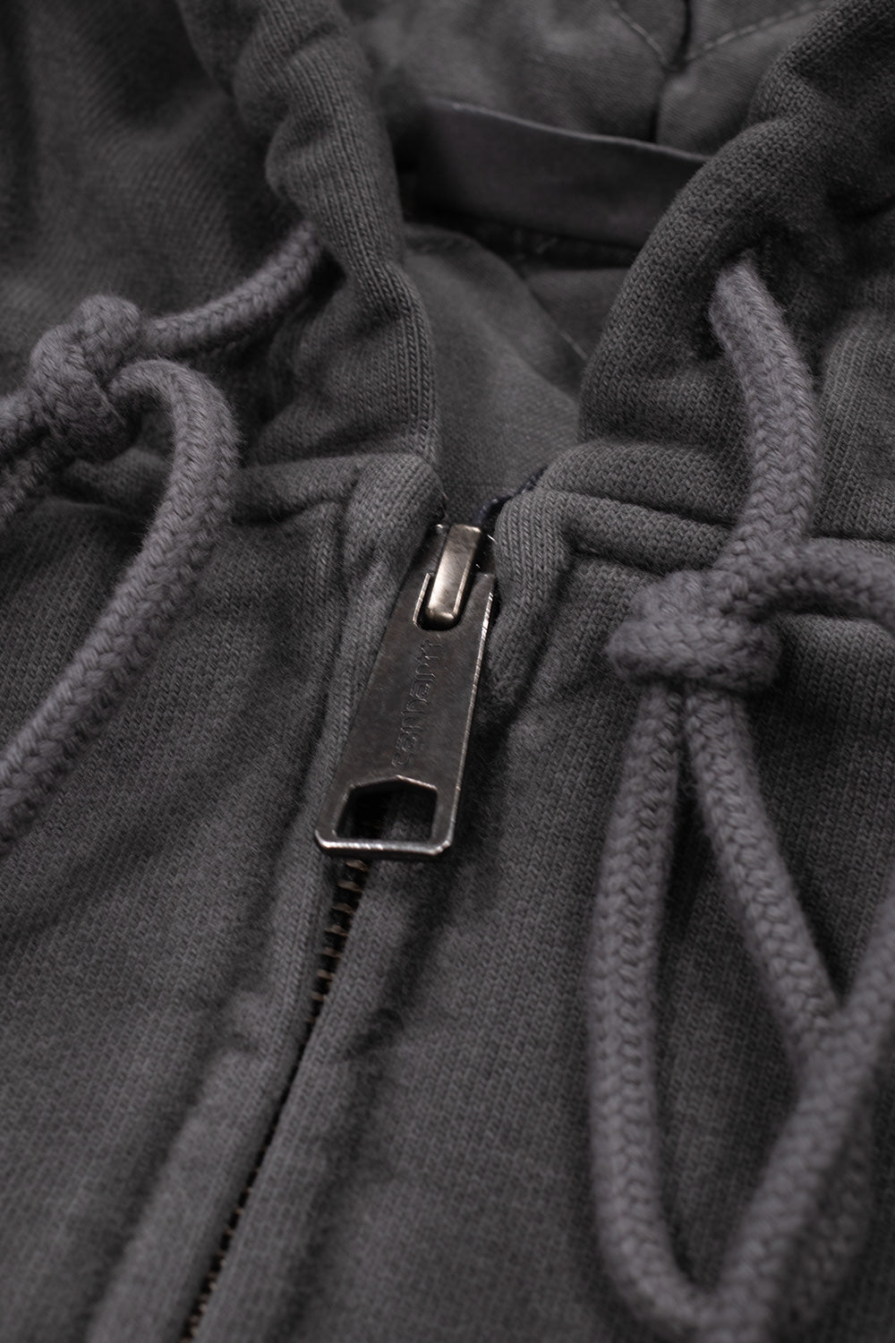 Macro close-up shot of the front neckline on the Carhartt WIP Hooded Vista Jacket, highlighting the antique silver metallic zipper pull embossed with Carhartt block lettering, heavy-duty zipper teeth, and knotted matching dark grey-black round drawstrings.