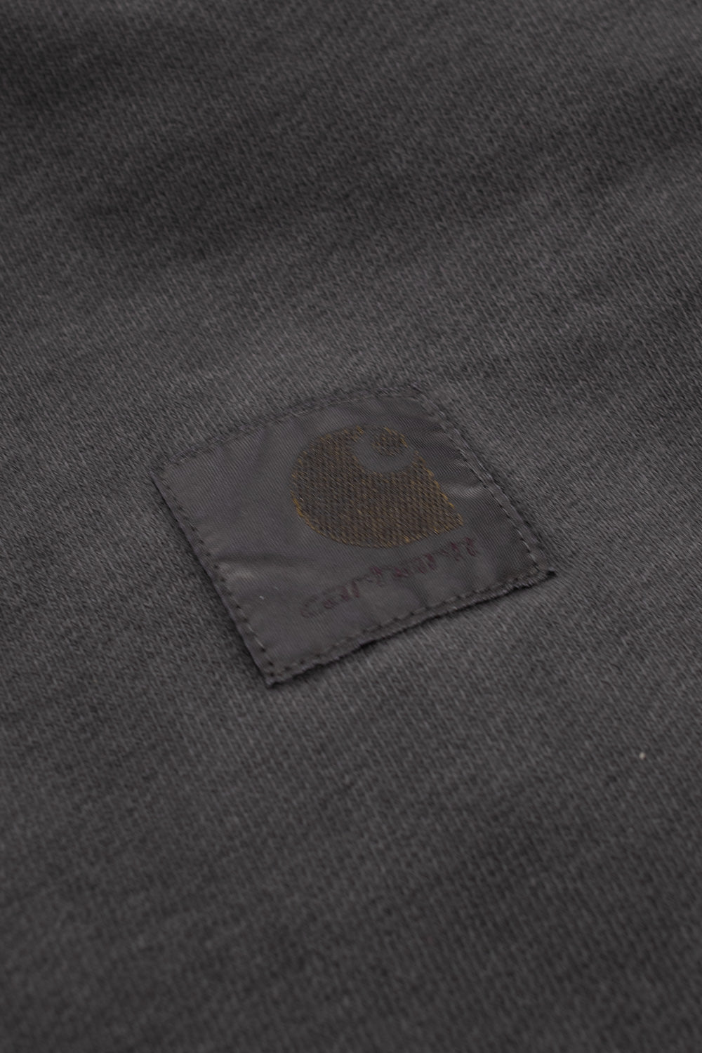 Detailed studio close-up of the tonal square branding patch on the washed black Carhartt WIP Hooded Vista Jacket, showing the subtle gold-and-brown woven "C" logo and muted lettering stitched onto the garment-dyed cotton fleece.