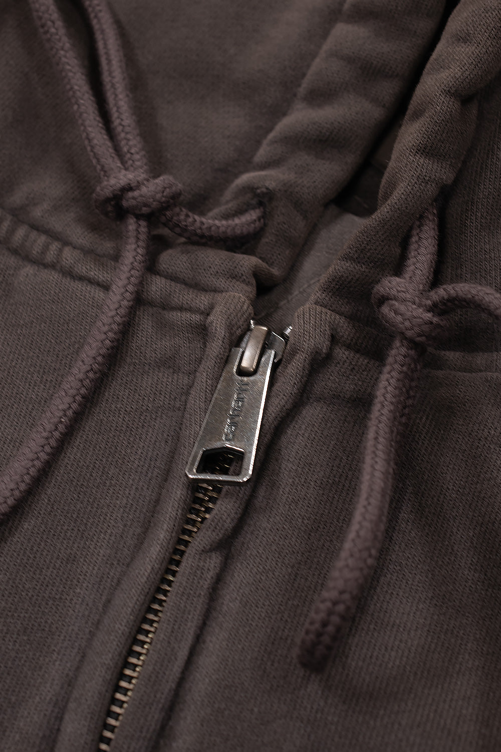 Macro close-up view of the front zipper closure on the Carhartt WIP Hooded Vista Jacket, highlighting the antique silver metal pull tab engraved with Carhartt branding, heavy zipper teeth, and knotted matching tobacco brown hood drawstrings.