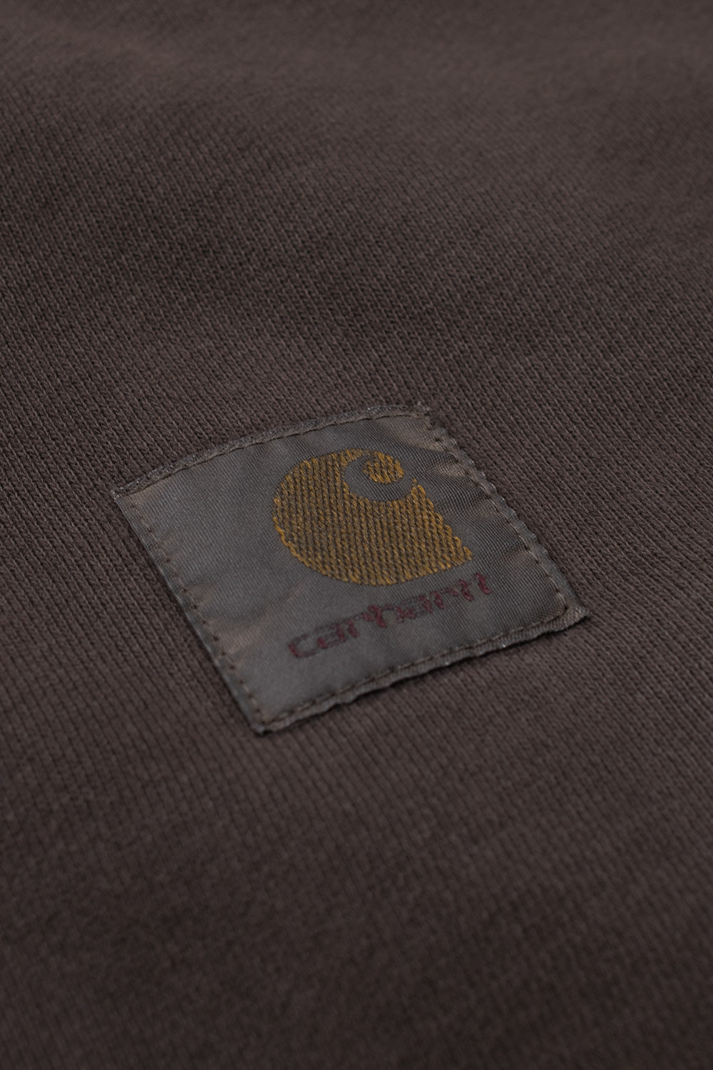Detailed studio close-up of the tonal square brand patch on the chest of the Carhartt WIP Hooded Vista Jacket, displaying the woven golden-brown "C" logo and muted maroon Carhartt script stitched onto washed tobacco garment-dyed cotton fleece.