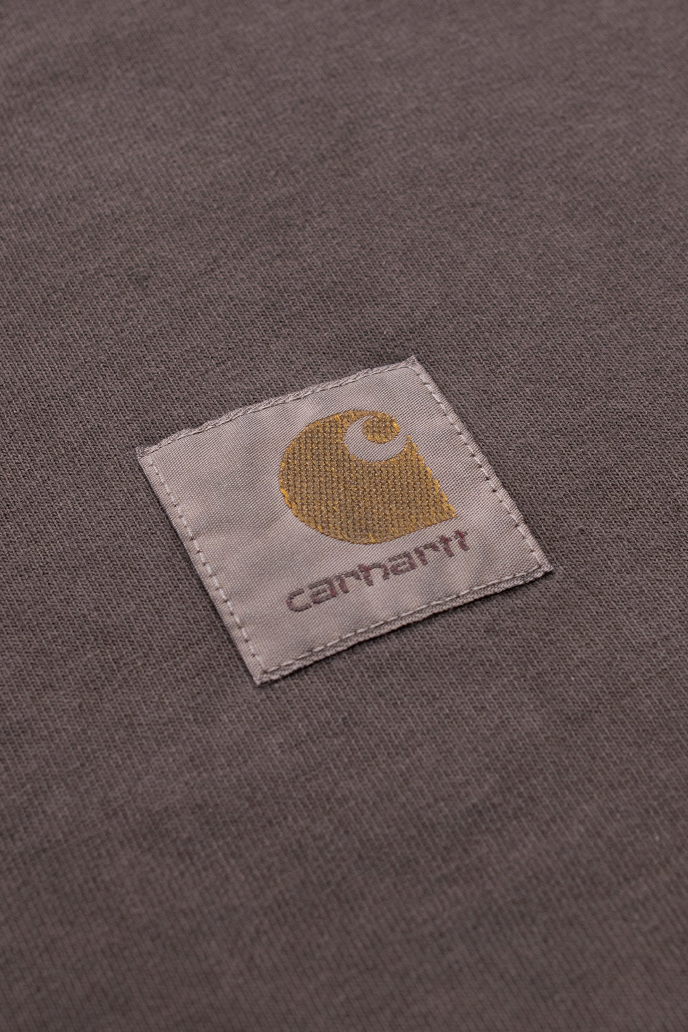 Detailed macro close-up of the washed cotton jersey fabric on the Carhartt WIP L/S Vista T-Shirt in Tobacco, highlighting the fine knit texture and the signature square label patch with the golden-brown Carhartt 'C' wave logo.