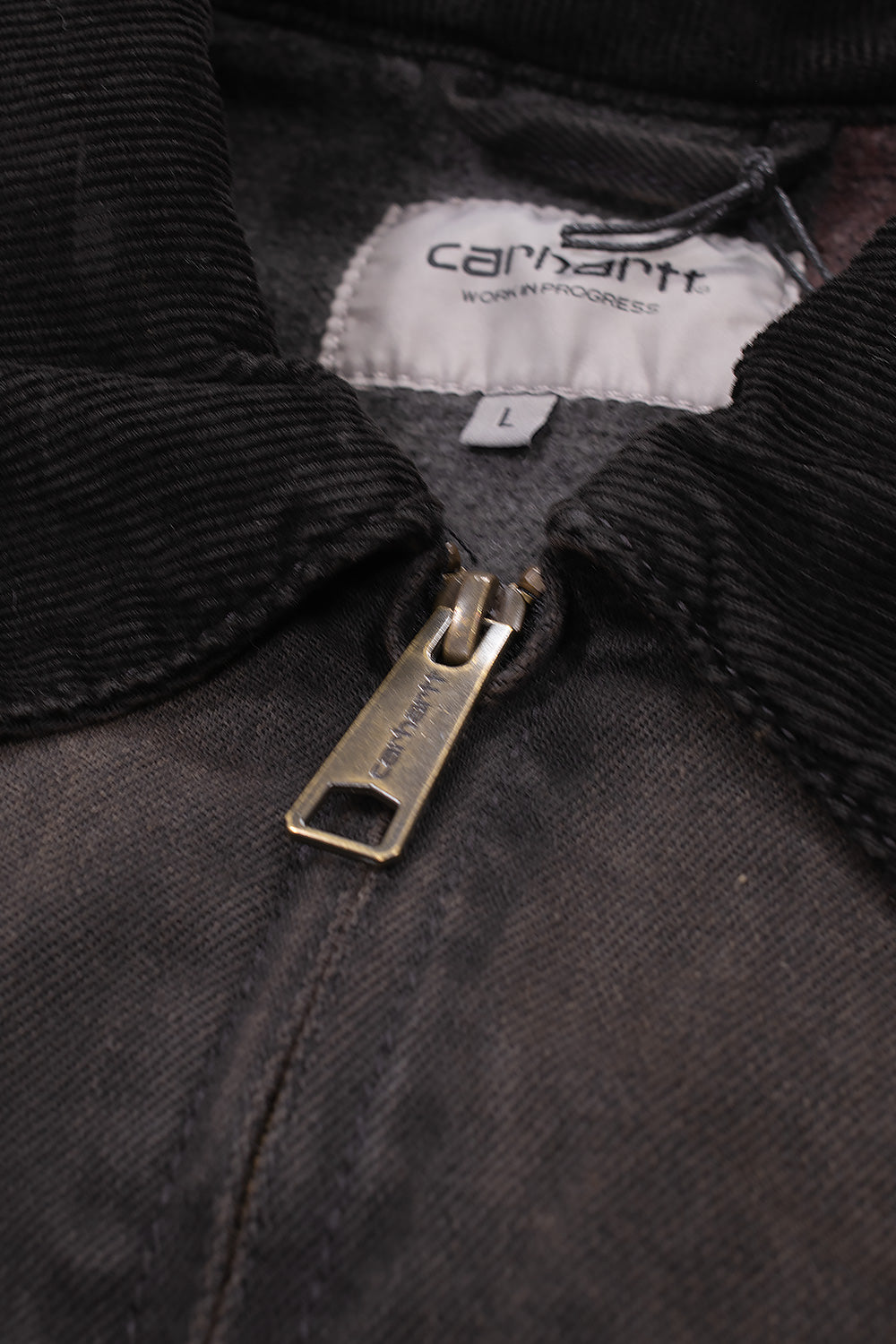 Macro close-up shot of the neckline on the Carhartt WIP OG Detroit Jacket, detailing the fine-ribbed dark brown corduroy collar, antique brass zipper pull engraved with Carhartt script, and internal white woven "Carhartt Work In Progress" collar label.