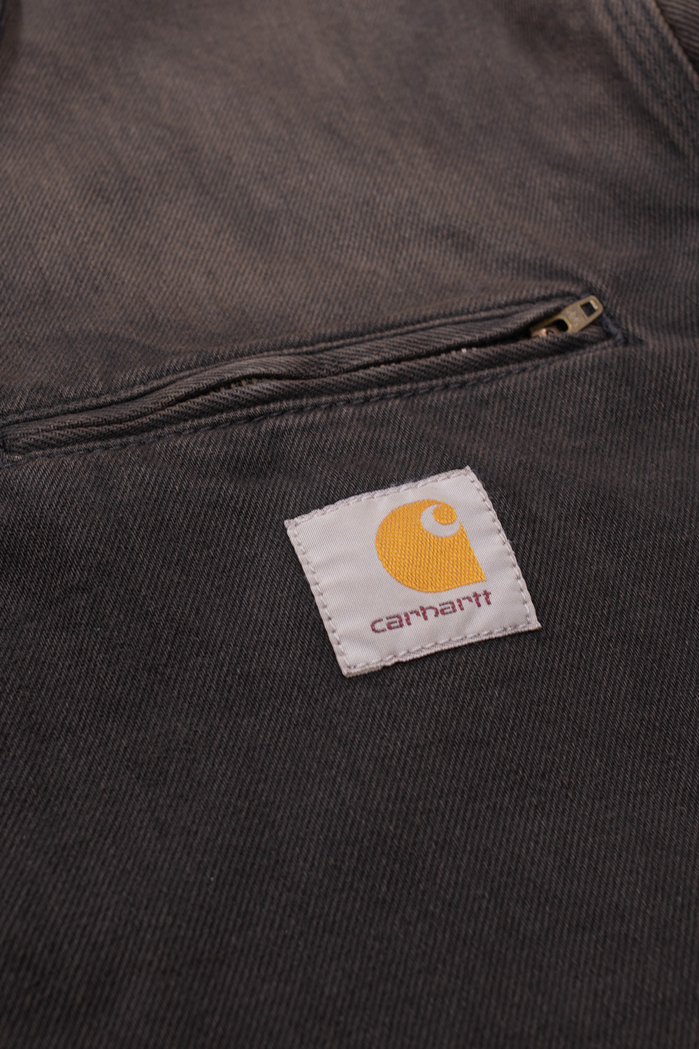 Detailed studio close-up of the chest area on the Carhartt WIP OG Detroit Jacket, showing the horizontal welt pocket with brass zipper closure above the iconic white woven square patch featuring the golden-yellow "C" emblem and brown script logo on faded black-brown denim.
