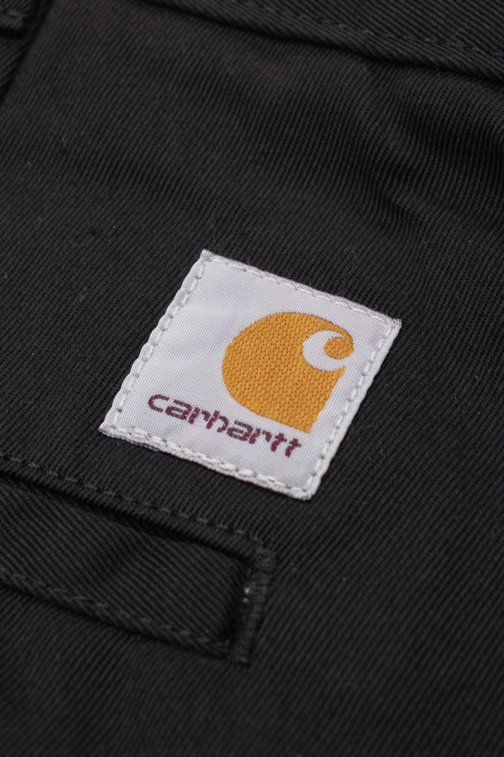 Macro close-up shot of the square woven Carhartt brand patch stitched above the back right welt pocket on the black rinsed OG Master Pant, showing the yellow Carhartt C logo emblem and burgundy lettering over black diagonal twill fabric.