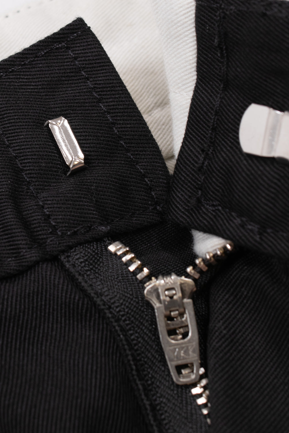 Detailed view of the front waistband closure on the Carhartt WIP OG Master Pant, highlighting the hidden metal slide hook and eye fastener, silver YKK metal zipper pull, and off-white interior waistband lining.
