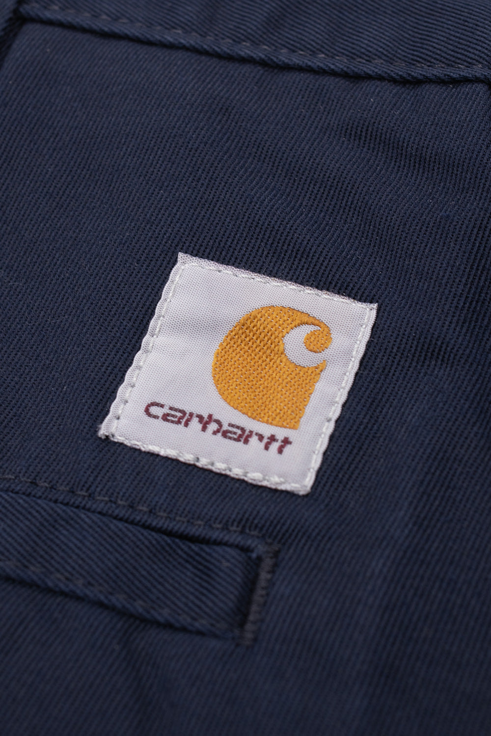 Macro close-up shot of the square woven Carhartt logo label stitched above the back right welt pocket on the dark navy blue OG Master Pant, showing the golden-yellow Carhartt C emblem and burgundy brand lettering over diagonal twill workwear fabric.