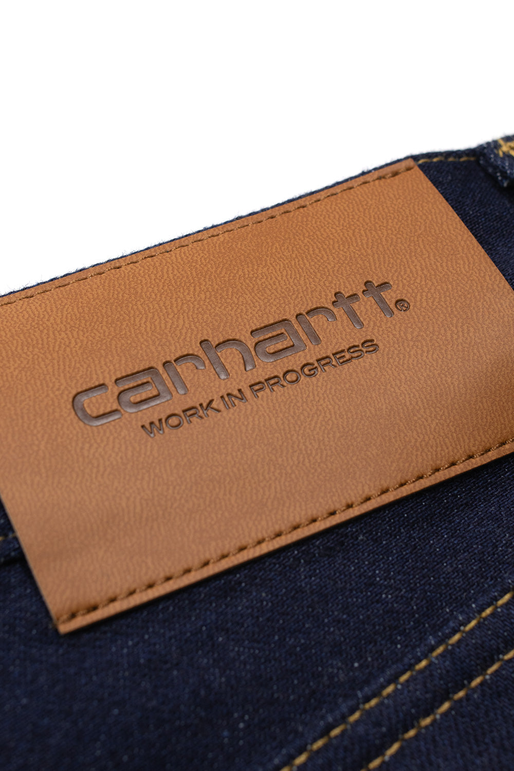 Macro close-up shot of the rear right waistband on the Blue Rigid Carhartt WIP Payson Pant, showcasing the rich tan leather patch debossed with the Carhartt Work In Progress wordmark logo beside clean double-needle contrast yellow stitching on raw dark blue denim.