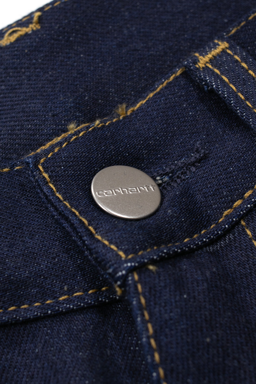 Detailed shot of the front waist closure on the Carhartt WIP Payson Pant, highlighting the metallic silver tack button engraved with Carhartt lettering set into durable raw dark indigo blue twill denim beside contrast stitching.