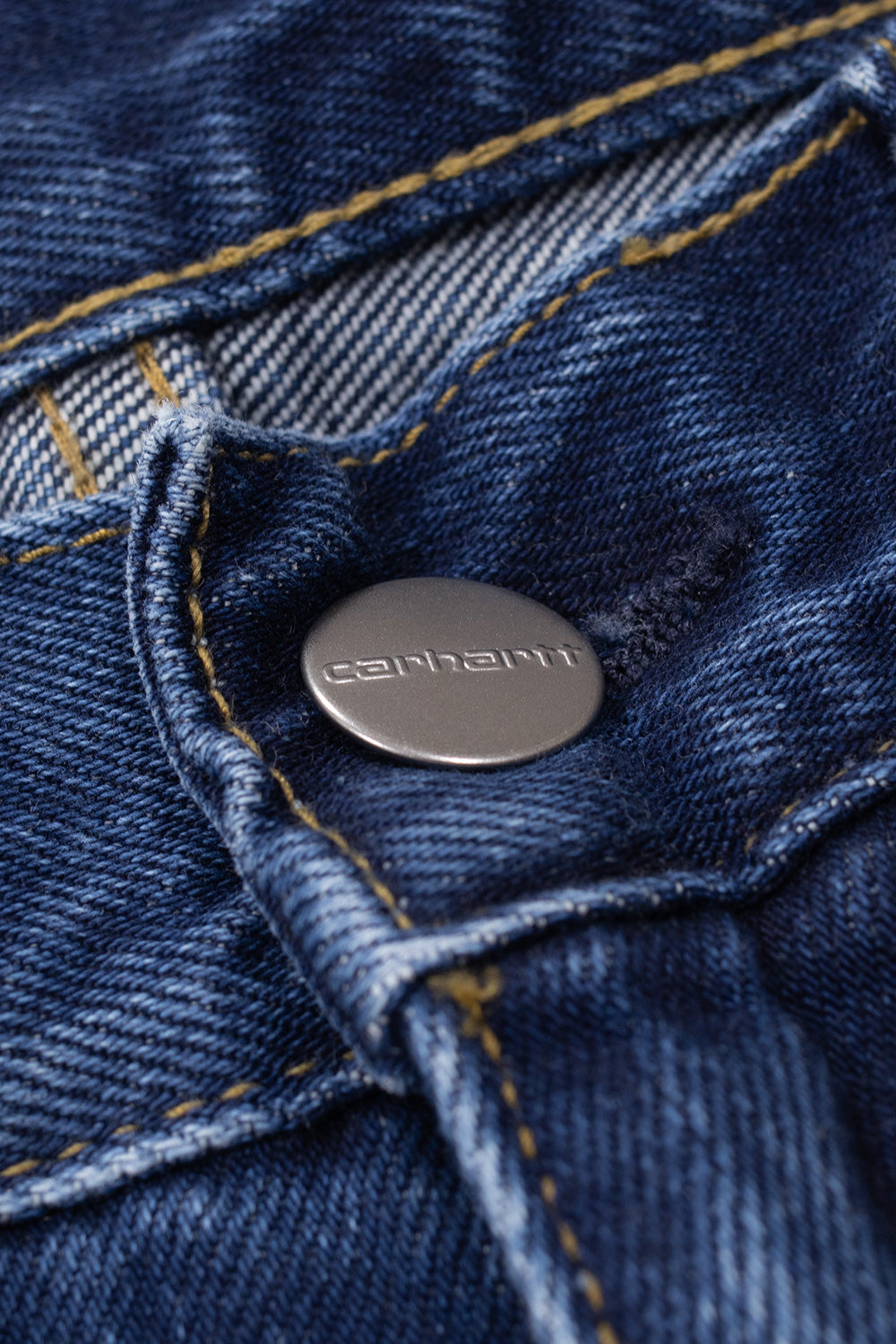 Detailed view of the front waist closure on the Carhartt WIP Payson Pant Blue Stone Washed, showcasing the engraved metallic silver shank button with Carhartt lettering set into soft stone-washed blue denim beside reinforced buttonhole stitching.