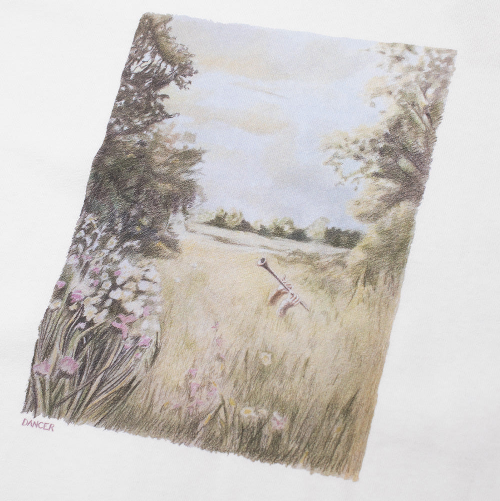 Detailed macro studio close-up of the chest graphic print on the Dancer Flute T-Shirt, highlighting the textured colored-pencil style pastoral artwork showing a scenic countryside meadow with trees, wild grass, wildflowers, and a flute in the foreground, finished with a subtle "DANCER" signature at the lower left corner