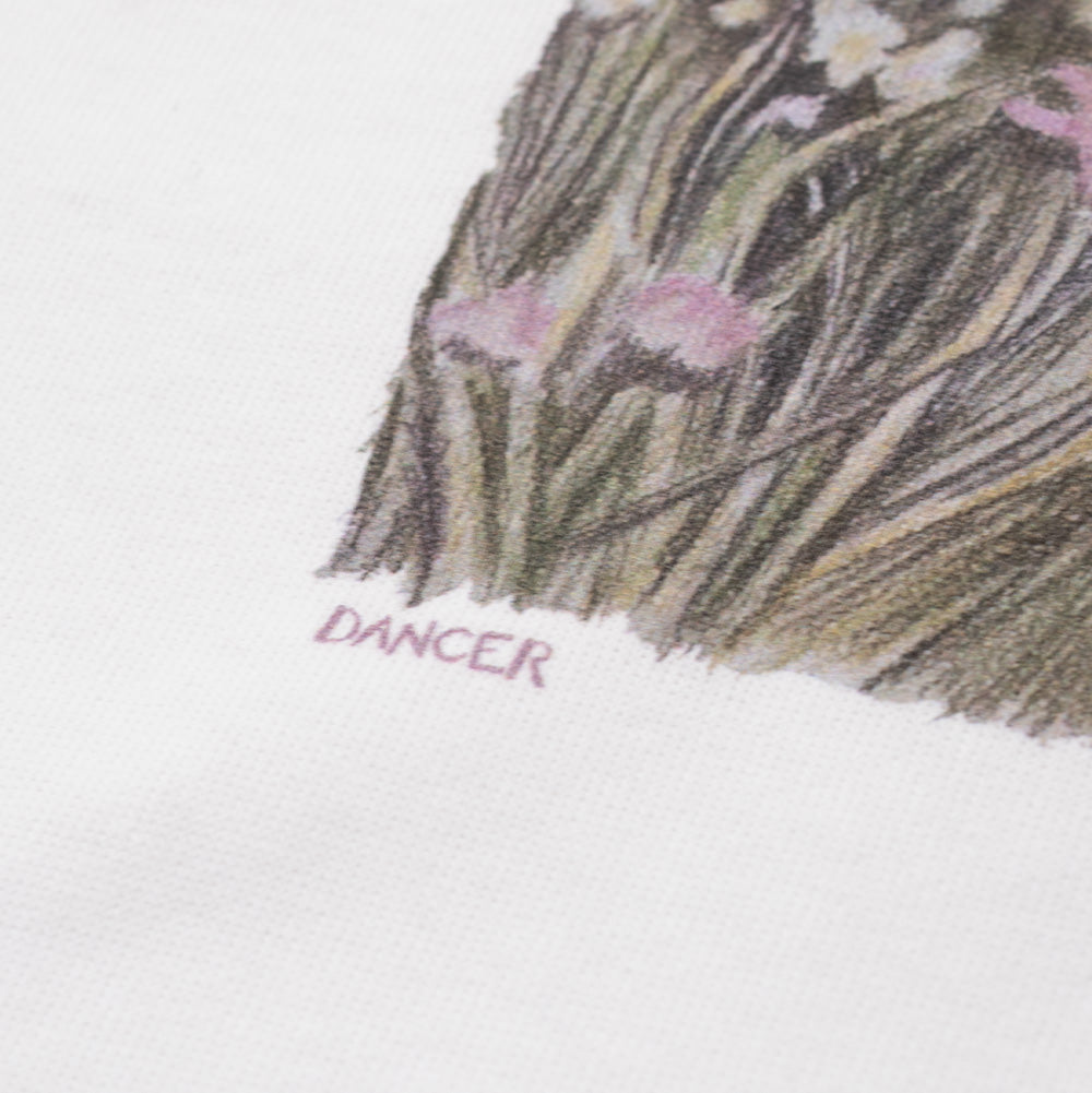 Extreme macro close-up of the lower left edge of the chest graphic on the Dancer Flute T-Shirt, focusing on the detailed sketch texture of pink meadow wildflowers and dry grass clumps next to the discreetly printed "DANCER" logo typography on smooth white cotton jersey fabric.