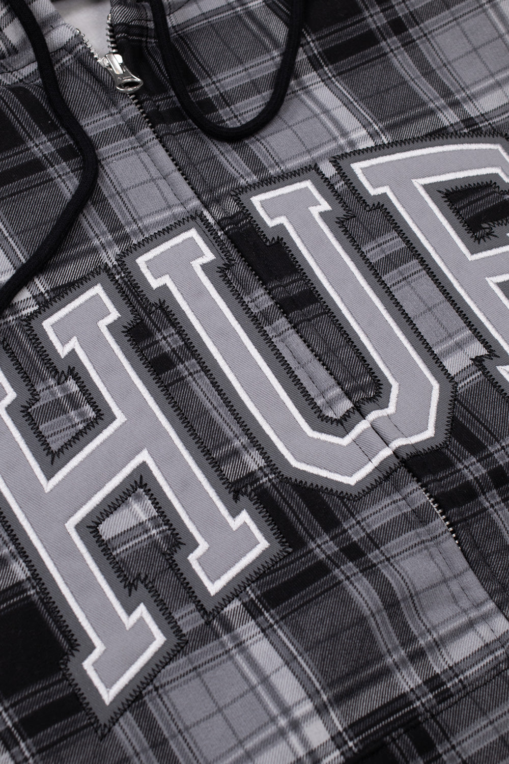 Detailed angled close-up of the collegiate logo patch on the chest of the black HUF Arch Plaid hoodie, highlighting the grey tackle twill letters with white borders and black zig-zag embroidery stitching set against the black and white cotton flannel plaid fabric, beside the silver YKK zipper teeth and black drawstrings.