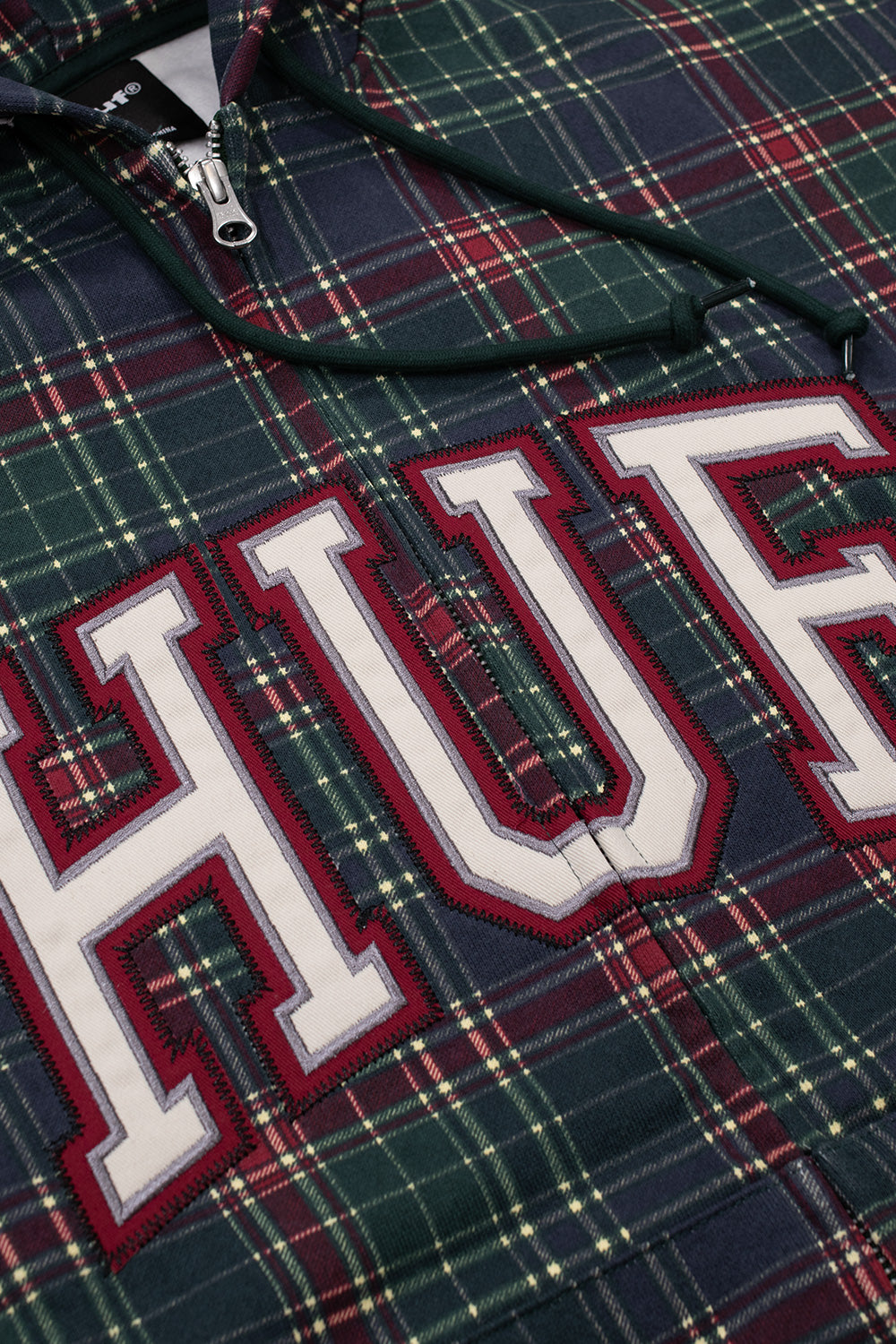 Detailed angled close-up of the varsity logo applique across the center chest of the HUF Arch Plaid hoodie, highlighting the layered white tackle twill letters with dark red outline embroidery and zig-zag stitching set over the green and red cotton plaid pattern beside the metallic YKK zipper track and dark green drawstrings.