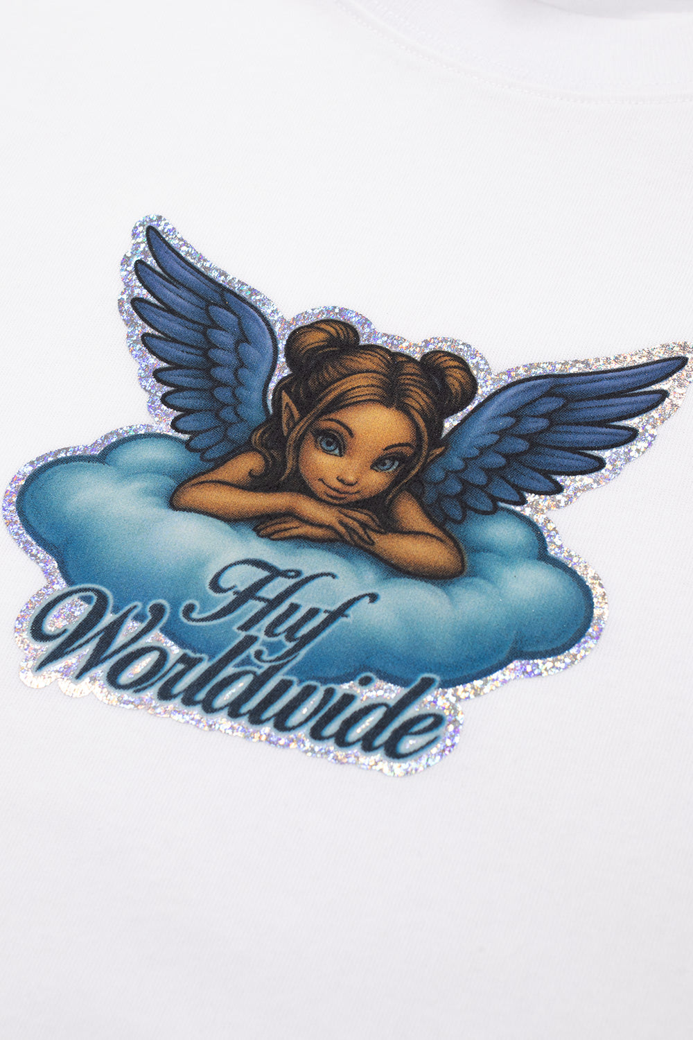 Macro close-up shot of the front chest graphic on the white HUF Bliss T-Shirt, highlighting the detailed illustration of a winged angel with blue feathers resting on a pastel cloud, dark script "Huf Worldwide" lettering, and the shimmering metallic silver glitter outline running along the edge of the artwork on white cotton fabric