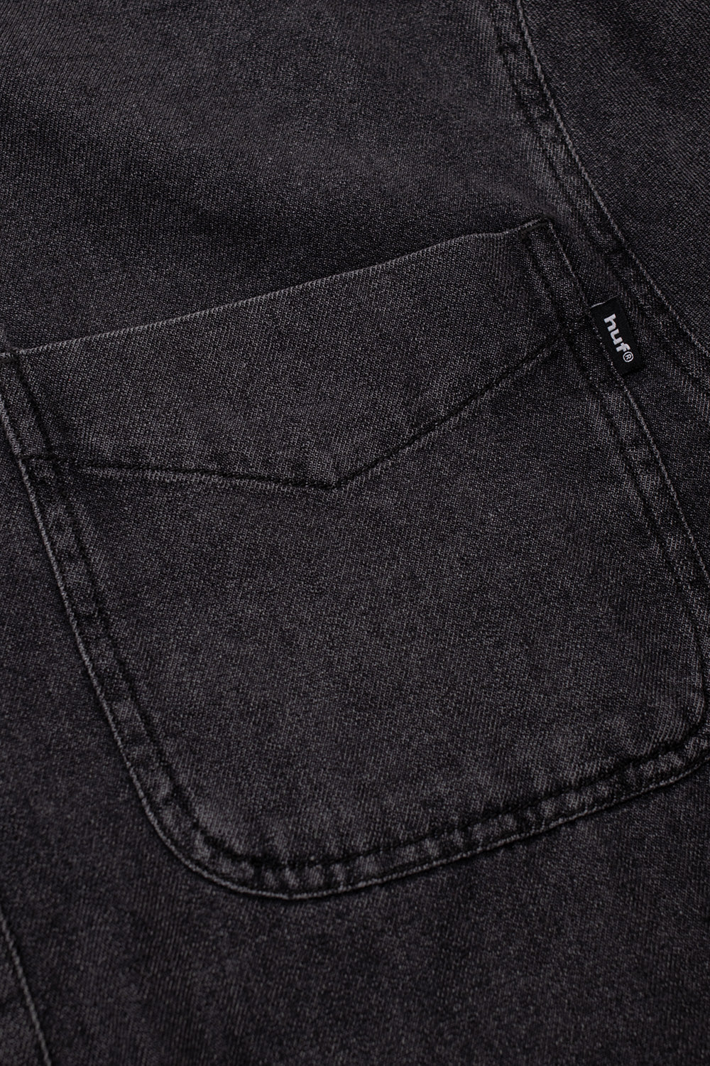Close-up view of the single chest patch pocket on the black HUF Carson Overdyed Denim Shirt, showing the top-stitched curved lower corners, horizontal reinforcement bar tacks, and the subtle black woven HUF brand flag label attached to the outer edge