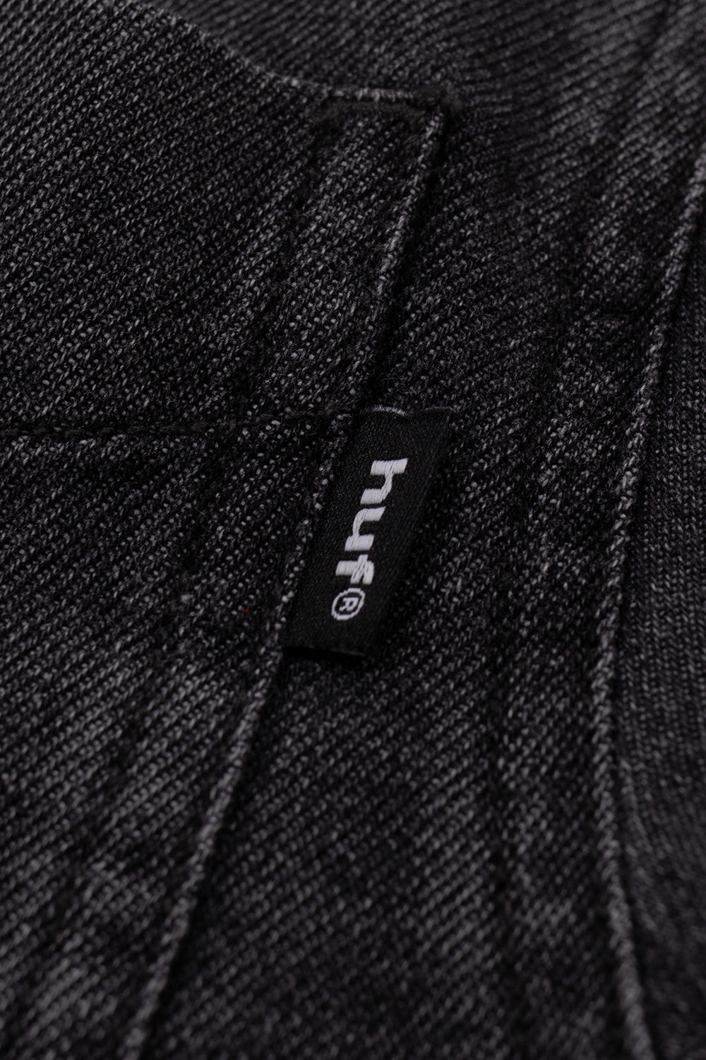 Macro detail of the woven brand flag label on the chest pocket of the HUF Carson Denim Shirt, displaying crisp white embroidered "huf" lettering with a registered trademark symbol against a black woven tag, framed by the textured black faded denim weave