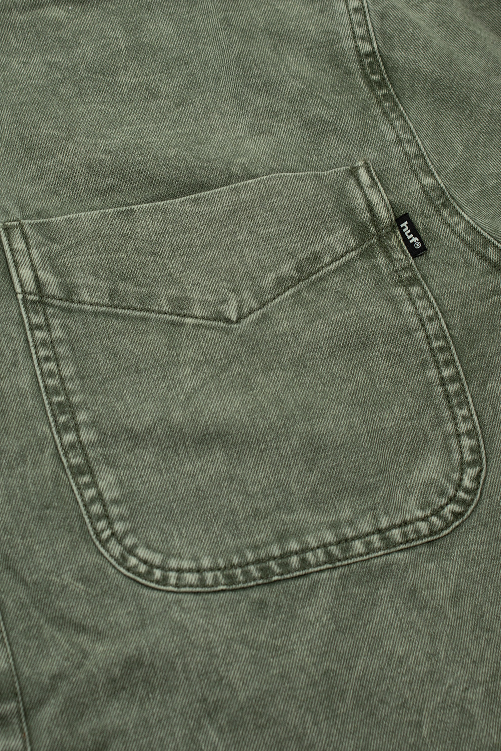 Close-up view of the open chest patch pocket on the olive HUF Carson Overdyed Denim Shirt, showing the angled internal pen-stitch divider line, reinforced top corners, and the black woven HUF brand flag label sewn along the right vertical seam