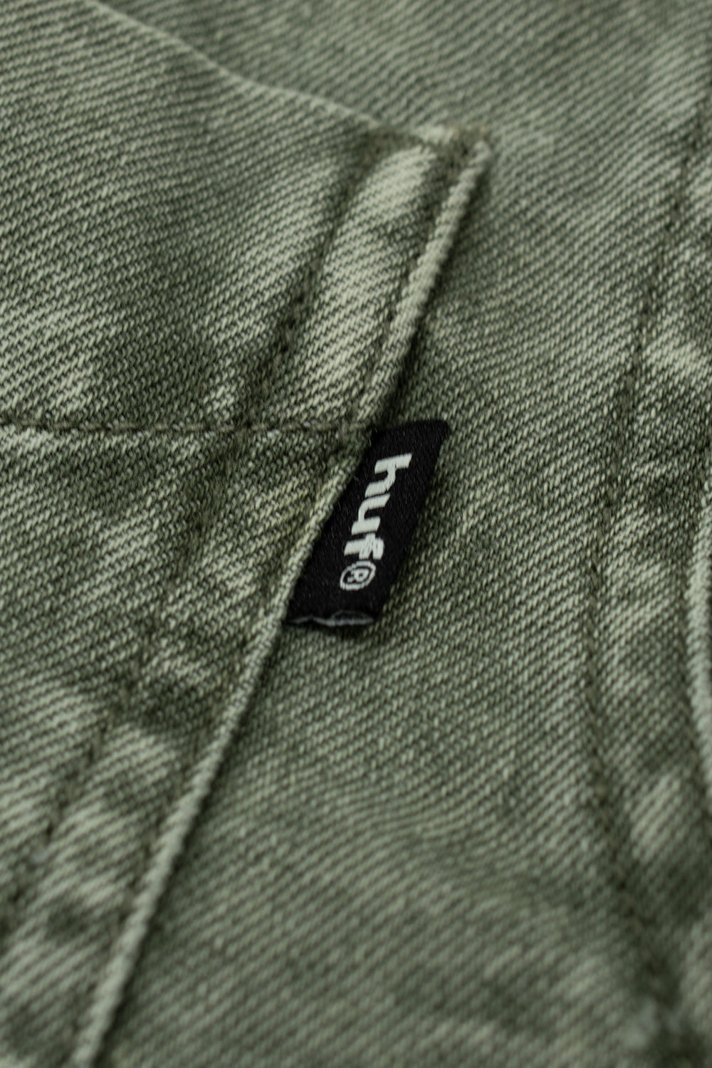 Macro detail of the black woven HUF logo flag label on the chest pocket of the olive Carson Denim Shirt, displaying crisp white embroidered "huf" lettering with a registered trademark symbol against the heavily washed olive-drab cotton denim grain